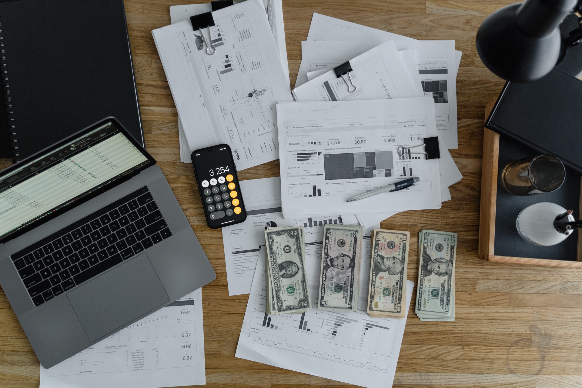 Zdjęcie biurka, na którym leżą pieniądze, papiery, laptop i telefon z uruchomionym kalkulatorem
